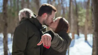 Couple kissing while holding an engagement ring toward the camera