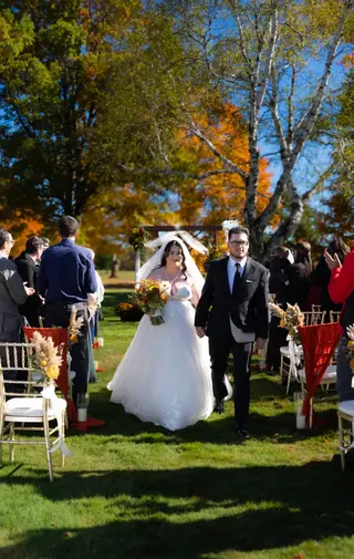 Bride walking along a grassy ceremony aisle beside a suited companion
